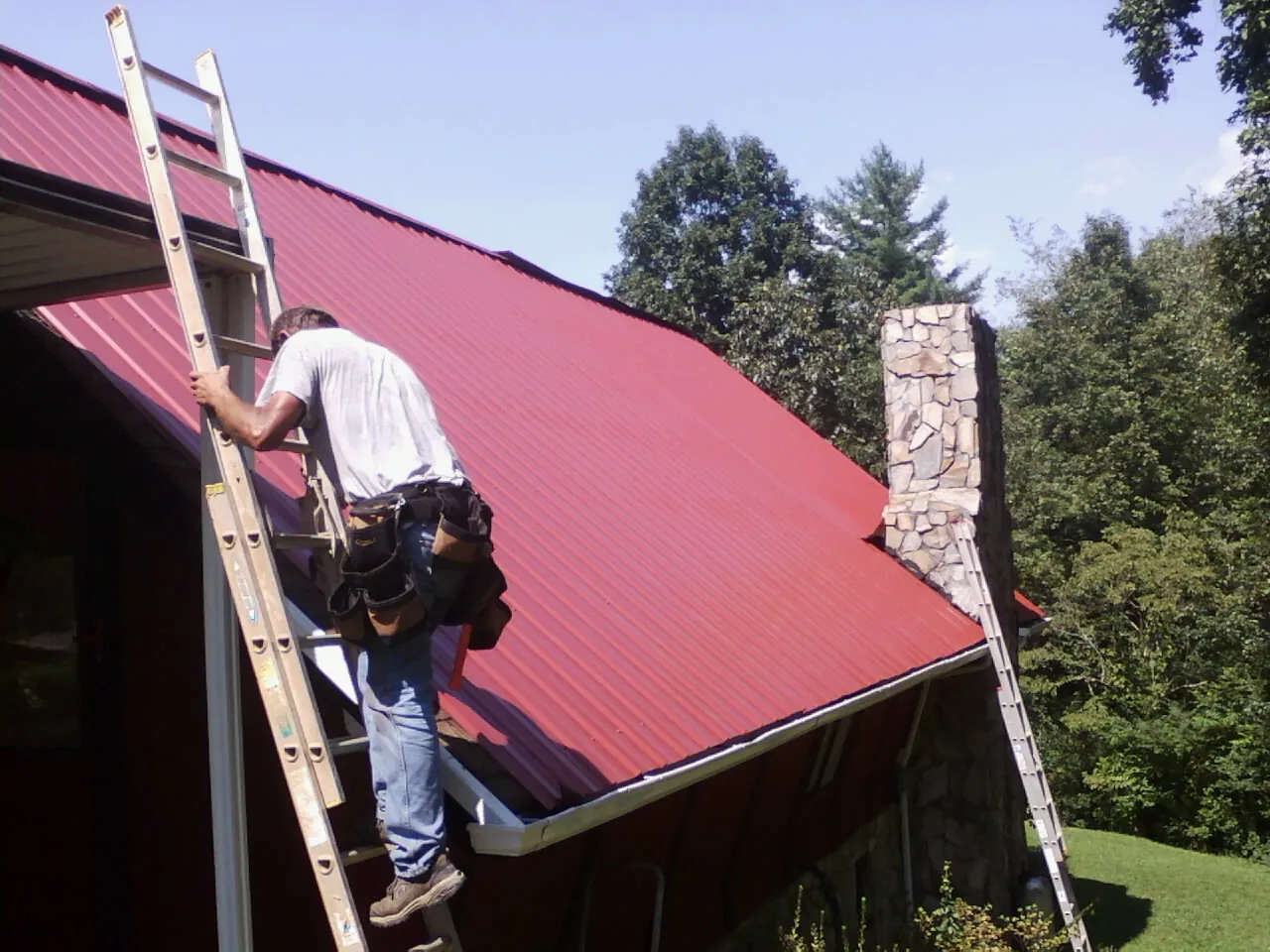 Roofer on a ladder beside a red metal roof and stone chimney — Soffit & Fascia Repair by Roofing Brothers in Belfry